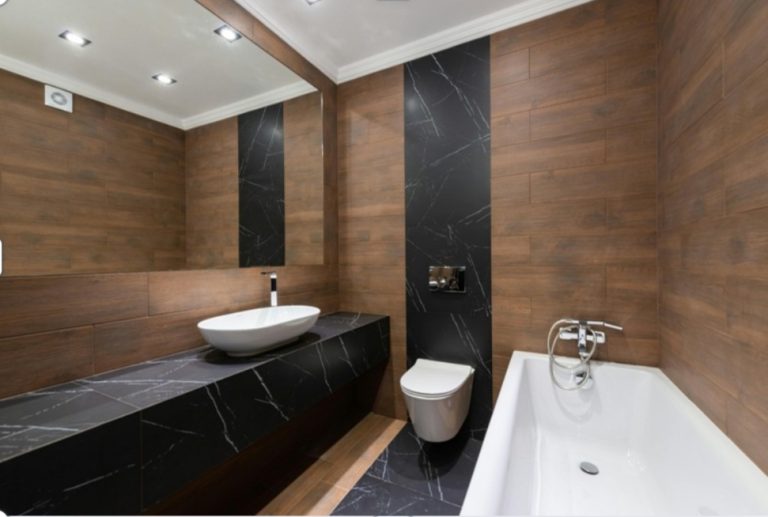 Une salle de bain élégante avec des murs revêtus de bois sombre et des accents de marbre noir. On y voit un lavabo vasque blanc posé sur un plan de travail noir, un WC suspendu et une baignoire blanche sur la droite.