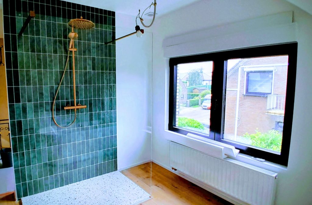 Une salle de bain contemporaine avec une douche à l'italienne dotée d'un mur en carrelage vertical vert émeraude et d'une robinetterie dorée. Le sol de la douche est en terrazzo clair. À côté, une grande fenêtre à cadre noir donne sur l'extérieur, avec un radiateur blanc installé dessous.