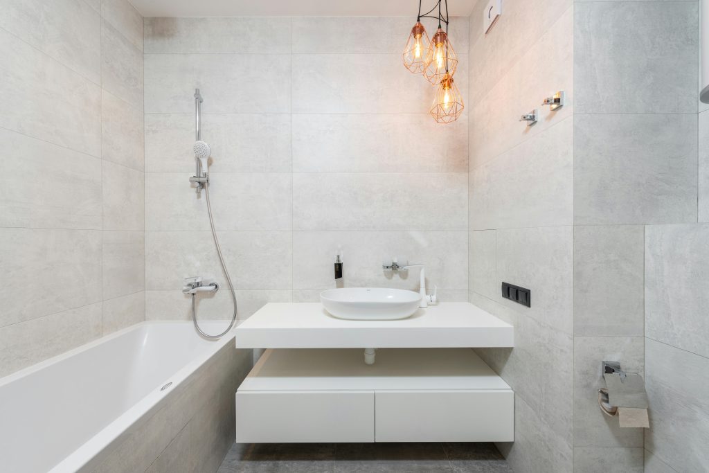 Une salle de bain lumineuse avec des carreaux gris clair. Elle comprend une baignoire blanche sur la gauche et un meuble vasque blanc suspendu au centre. Trois lampes suspendues de style industriel avec des ampoules à filament apportent une touche chaleureuse.