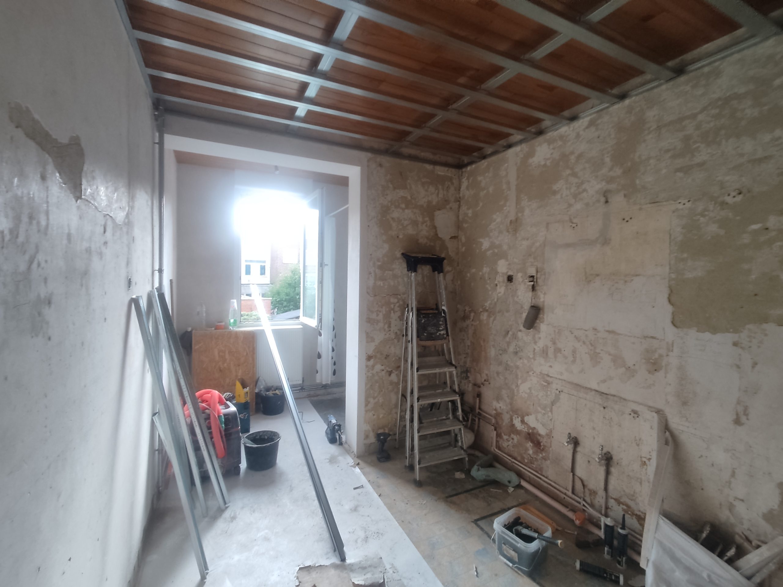 Une pièce en plein chantier de rénovation. Les murs sont en pierre et brique apparente avec des traces d'ancien enduit. Le plafond montre une structure de rails métalliques pour un futur faux plafond sous des solives en bois. Une échelle et des outils de construction sont visibles au centre.
