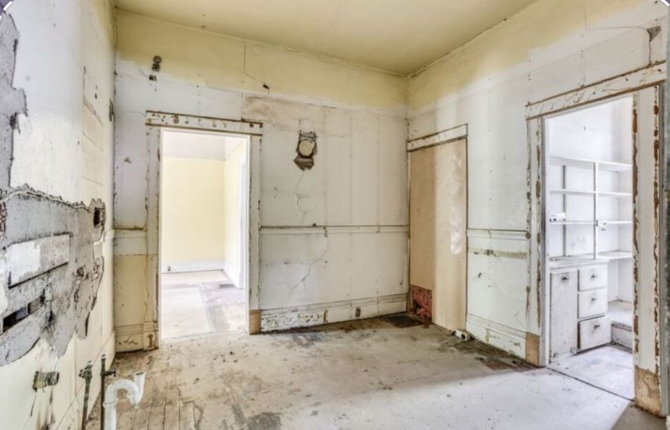 Une ancienne cuisine complètement vidée avant rénovation. Les murs présentent des traces de vieux revêtements arrachés, et le sol est en béton brut. On aperçoit deux ouvertures de portes menant vers d'autres pièces vides.