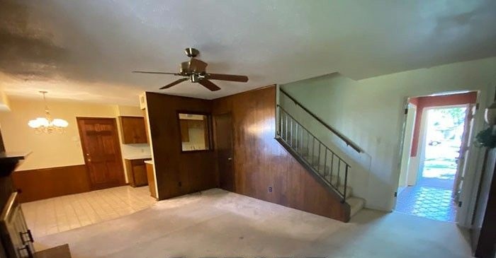 Vue d'un salon spacieux avant rénovation avec une moquette beige, des murs clairs et un grand ventilateur de plafond en bois. Un mur partiel recouvert de lambris en bois sombre sépare la pièce d'un escalier. Au fond, on aperçoit une porte d'entrée en bois et un coin repas.