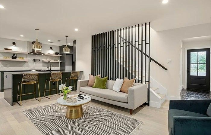 Un salon moderne à aire ouverte avec un canapé gris clair décoré de coussins verts et orange. Derrière le canapé se trouve une cloison décorative à lattes verticales noires masquant un escalier. À gauche, une cuisine moderne avec un îlot vert foncé et des tabourets dorés.