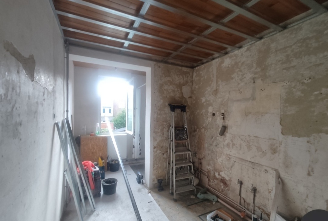 Une pièce en cours de rénovation avec des murs en pierre brute et un plafond dont la structure métallique est apparente sous des poutres en bois. On y voit une échelle double, des outils de chantier et des seaux au sol près d'une fenêtre ouverte.